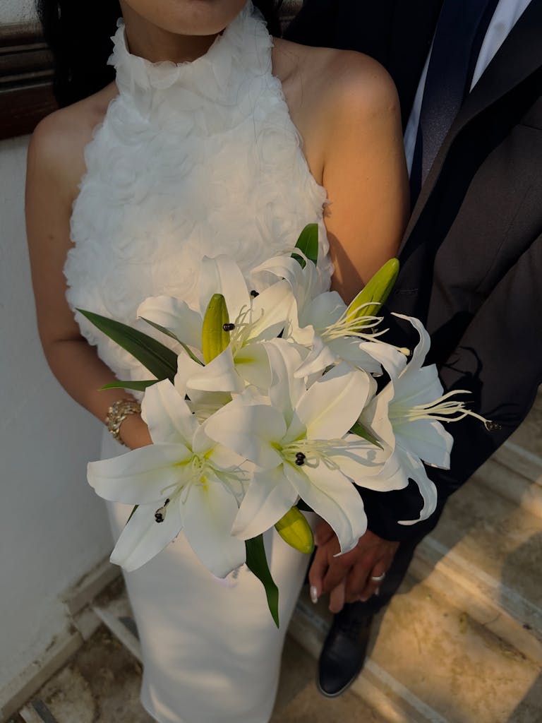 Bride holding a beautiful white lily bouquet in a close embrace with groom, showcasing elegant wedding attire.