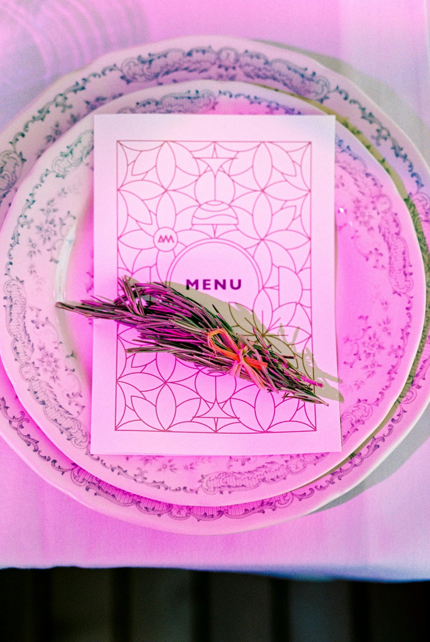 Stylish table setting featuring a menu card with floral arrangement on ornate plates, bathed in pink light.